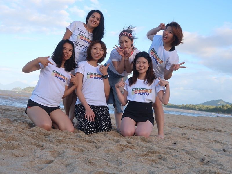 grupo de 6 chicas posando en la arena y detrás el mar. Llevan la misma camiseta blanca con un estampado de letras en medio